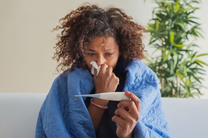 femme malade à la maison, enveloppée dans une couverture, avec une main tenant un mouchoir sur son nez et un thermomètre dans l'autre, se demandant si c'est la grippe, le covid ou le flurona